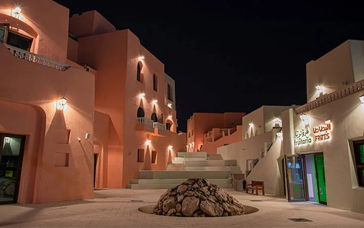 A nighttime view of tan, modern buildings with illuminated lights, surrounding a central courtyard that features a circular stone mound. A sign reading Furano Arts is visible on one building.