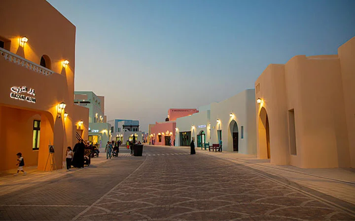 A wide, empty street lined with colorful, modern buildings featuring arched doorways and glowing lights at dusk, with a few people walking and soft evening sky overhead.