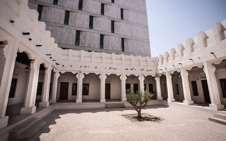 A courtyard with a single small tree in the center, surrounded by white traditional-style arches and columns, evokes Qatar heritage, with a modern multi-story building in the background under a clear sky—reminiscent of Msheireb Museums in Doha.