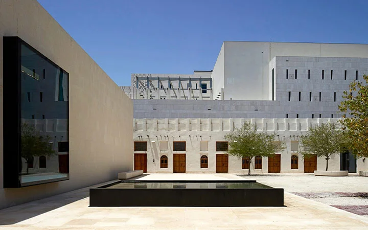 A modern courtyard at Msheireb Museums in Doha, Qatar, features minimalist architecture with light stone walls, a rectangular reflecting pool, neatly planted trees, and wooden doors under a clear blue sky.