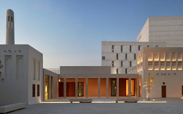 A modern building complex in Doha with minimalist beige facades, large windows, and a courtyard lit by warm lights at dusk. A slender tower and geometric structures, reminiscent of Msheireb Museums, stand against a clear sky.