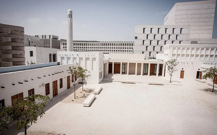 Modern and traditional white buildings surround a spacious, sandy courtyard with small trees and benches under a clear sky, showcasing the charm of Msheireb Museums—one of the leading Doha museums and Qatar cultural attractions.
