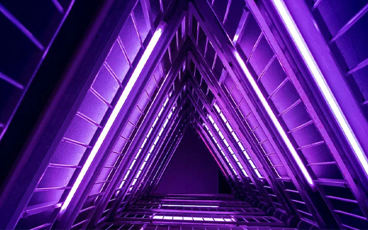 A triangular architectural structure at the Museum of Illusions is illuminated with vibrant purple and violet lights, creating a geometric, tunnel-like visual effect with repeating lines and glowing bars.