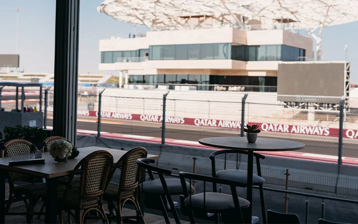 A view from a shaded seating area overlooking a racetrack, featuring empty tables and chairs in the foreground, with a modern building and Qatar Airways banners showcasing Premiere Hospitality Qatar in the background.