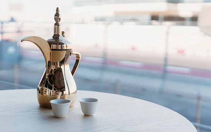 A traditional Arabic coffee pot and two small white cups sit on a light wooden table, reflecting the elegance of Premiere Hospitality Qatar against a blurred outdoor background.