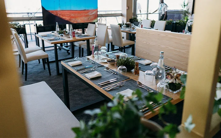 A modern restaurant dining area by Premiere Hospitality Qatar features tables neatly set with plates, glasses, cutlery, and water bottles. Succulent plants and flowers decorate the tables, while upholstered chairs and a large painting complete the setting.