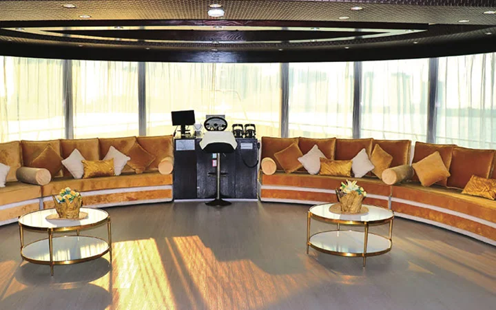 A spacious, sunlit lounge aboard a luxury houseboat in Qatar features curved golden sofas, round glass coffee tables with flower arrangements, and a ship’s control panel in the center against large windows with sheer curtains.