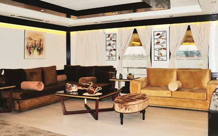 A modern living room aboard a Luxury Houseboat in Qatar, featuring brown velvet sofas, a round ottoman, wooden coffee tables, wall art, and large windows with white curtains overlooking a breathtaking waterfront view.