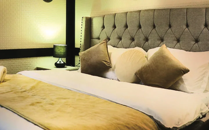A neatly made bed with a tufted gray headboard, white sheets, beige pillows, and a matching beige blanket creates a cozy retreat. Perfect for relaxing aboard a luxury houseboat in Qatar, the room features soft lighting and a sleek black bedside lamp.