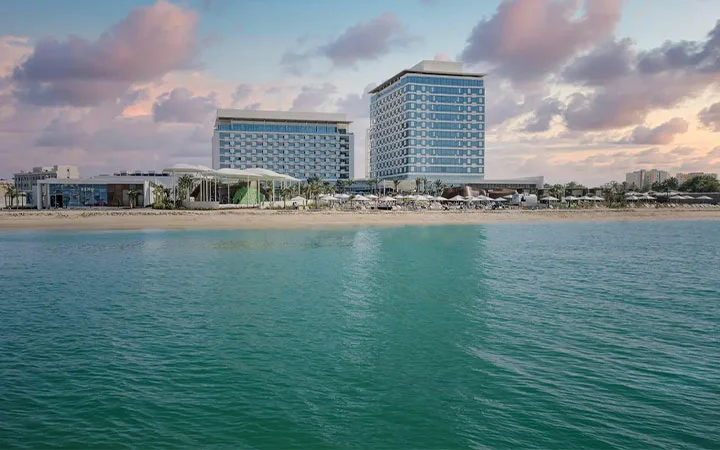 A modern beachfront hotel, Rixos Gulf Beach, with two large buildings stands along a sandy shore, fronted by calm turquoise water under a sky with scattered clouds at sunset.