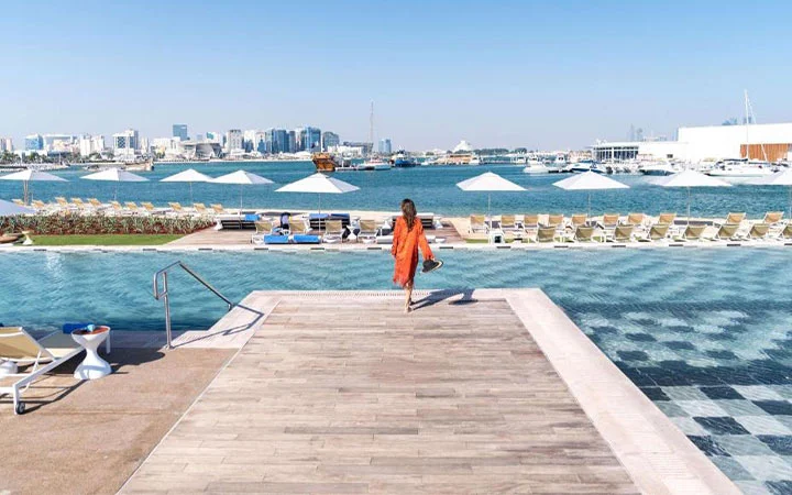 A woman in an orange dress walks toward a row of lounge chairs and umbrellas by a waterfront infinity pool at Rixos Gulf Beach, a luxury hotel with city buildings visible across the water under a clear blue sky.