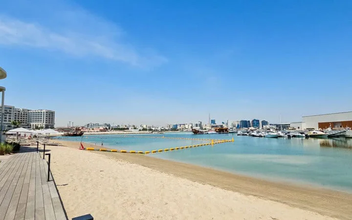 Sandy beach with calm, clear water; yellow floating barriers separate swimming area. Enjoy stunning views of modern buildings and yachts across the water, with Rixos Gulf Beach nearby under a bright blue sky.