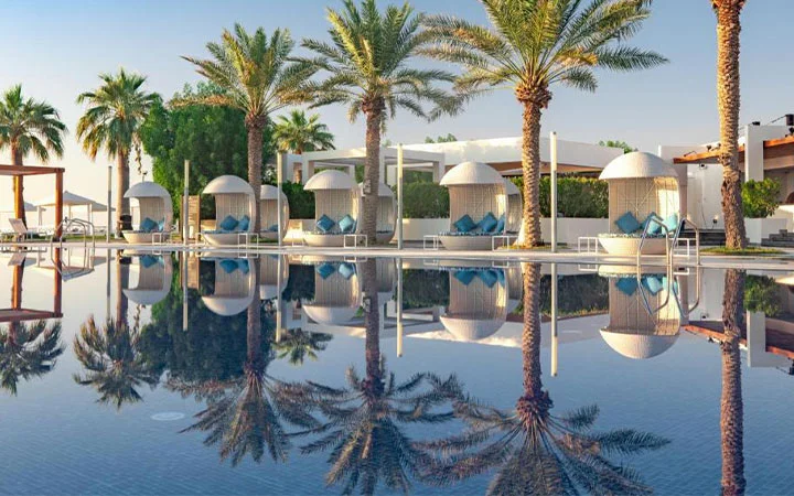 A luxurious outdoor pool at Sealine Beach Resort is lined with palm trees and modern cabanas with blue cushions, all reflected in the calm water under a clear sky.