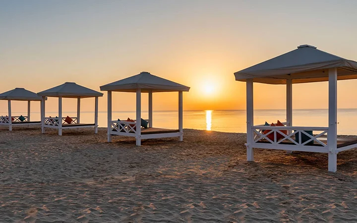 Four white beach cabanas with pillows stand on the sandy shore at sunrise, facing the calm sea and a clear sky. Soft light and reflections highlight the tranquil beauty of Sealine Beach Resort.
