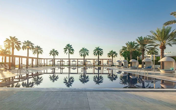 A luxurious outdoor pool at Sealine Beach Resort, surrounded by palm trees, cabanas, and lounge chairs, reflects the clear blue sky at sunrise or sunset, creating a tranquil and inviting beach resort atmosphere.