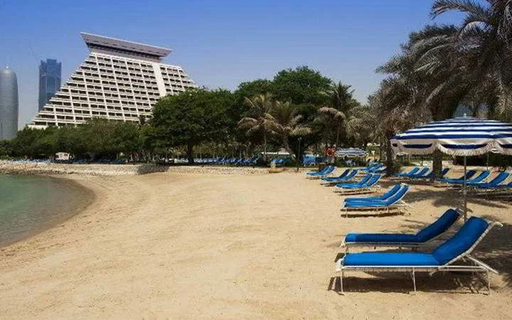 A sandy Doha Beach lined with blue lounge chairs and umbrellas, bordered by palm trees. In the background, the distinctive pyramid-shaped Sheraton Doha hotel and tall modern buildings rise under a clear blue sky.