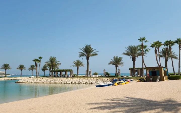 A sandy beach with clear, calm water at Sheraton Doha Beach is lined with palm trees and shaded cabanas. Several colorful kayaks and paddleboards are stacked near a hut on the shore under a clear blue sky.