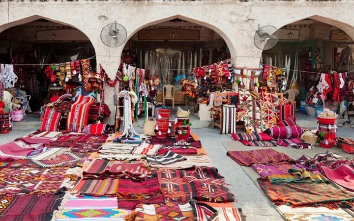 Colorful woven rugs, textiles, and cushions are displayed outside a market stall with arched entrances. Patterns are geometric and vibrant, and fans are mounted above the stalls.