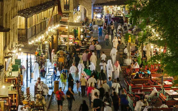 A bustling market street at night, filled with people walking and shopping, outdoor cafes with red seating, and shops displaying various goods under warm lights. Trees and old-style architecture add charm to the lively scene.
