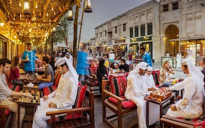 People sit at outdoor cafes in a bustling Middle Eastern market, some playing chess and others chatting. Traditional lanterns hang overhead, and historic buildings surround the lively scene at dusk.