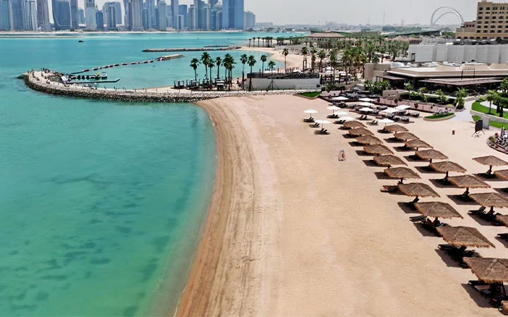 A sandy beach with thatched umbrellas and lounge chairs lines a turquoise shoreline at St Regis Beach. Palm trees and modern buildings rise in the background, with a city skyline across the water under a clear sky.