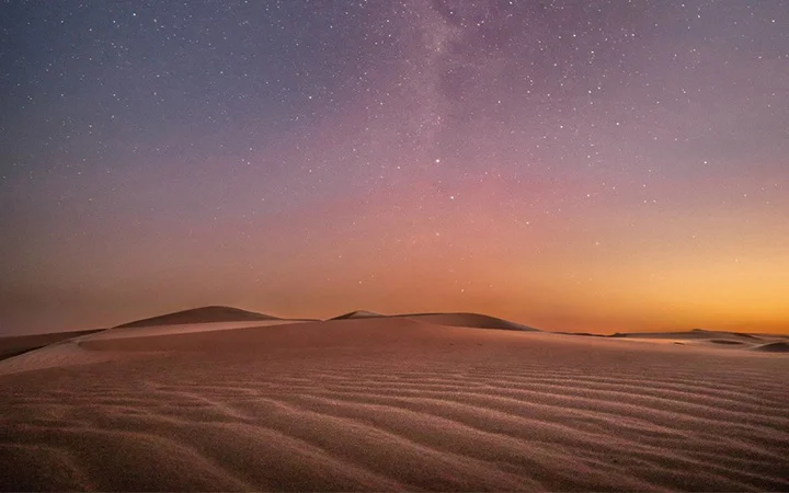 Rippling sand dunes under a twilight sky invite star gazing, as countless stars and a faint band of the Milky Way shine above, with purple and orange hues blending near the horizon—perfect for a magical desert adventure.