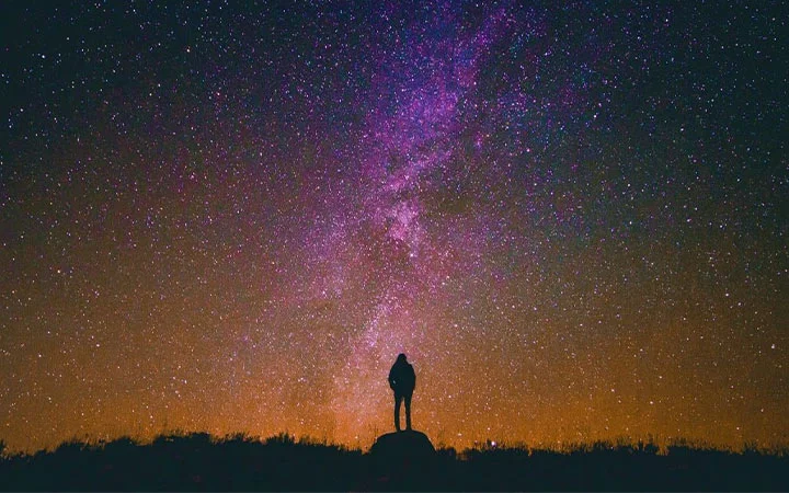 A person stands on a hill, silhouetted against a colorful night sky filled with stars and a visible stretch of the Milky Way, enjoying a moment of star gazing as orange light glows on the horizon.