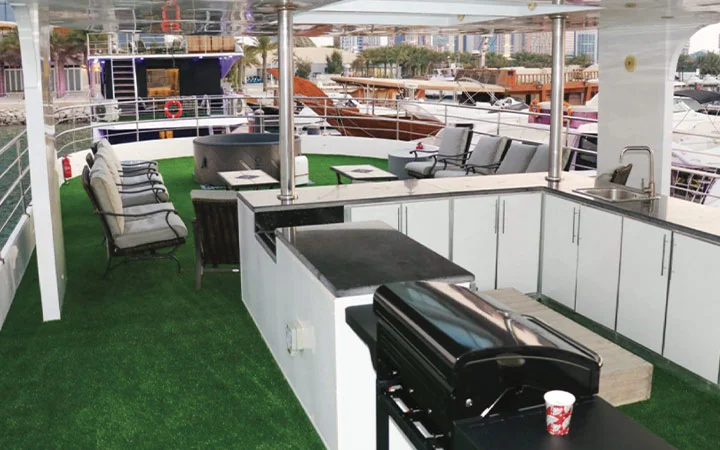 A Signature Private House Boat yacht deck with artificial grass flooring, outdoor seating, a counter with a barbecue grill, sink, and stool. Boats and buildings can be seen in the background near the water.