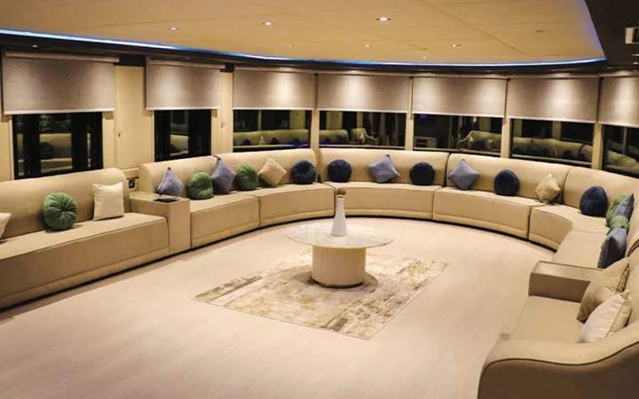 A spacious, modern lounge aboard a Signature Private House Boat features curved beige sofas, colorful pillows, large windows with shades, and a circular coffee table with a vase in the center on a light rug.