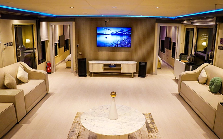 Modern, spacious living room in a Signature Private House Boat with neutral tones, two beige sofas, a round marble coffee table, and a large wall-mounted TV. Soft lighting and blue ceiling accent lights enhance the cozy atmosphere.