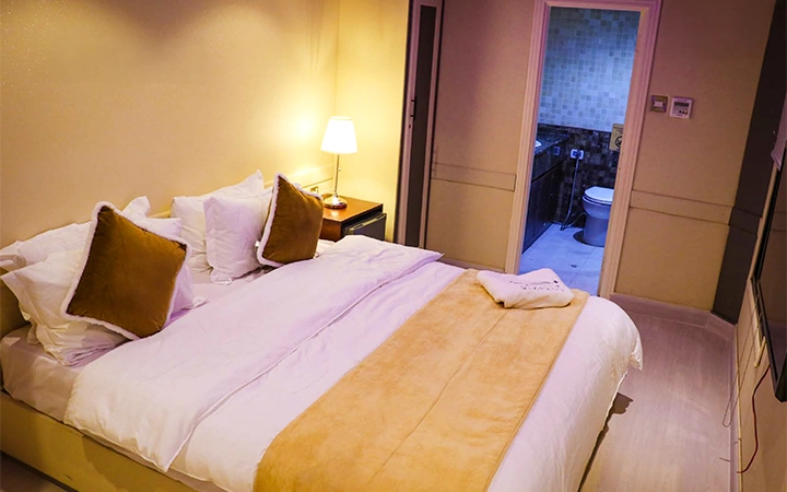 A cozy hotel room with a large bed, white sheets, and tan blankets, three brown pillows, and a bedside table with a lit lamp—just steps away from the bathroom—offers comfort reminiscent of a Signature Private House Boat retreat.