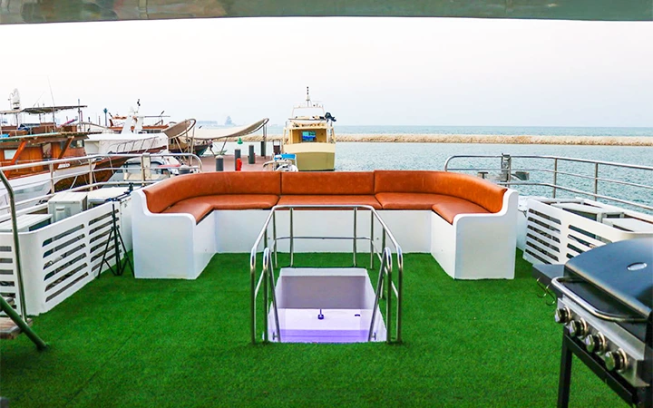 A Signature Private House Boat features a yacht deck with artificial green grass, an orange cushioned curved seating area, a small glass-covered opening, stainless steel railings, and a barbecue grill overlooking boats and the sea.