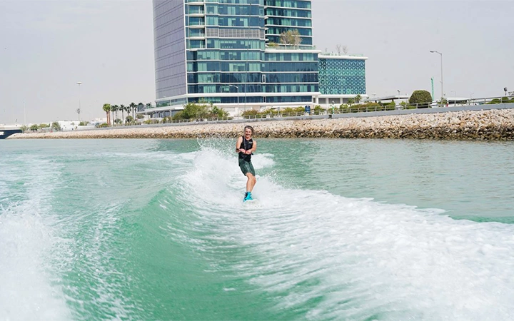 A person wakeboarding in Qatar on green water near a rocky shoreline, with a modern glass building and palm trees in the background under a clear sky.