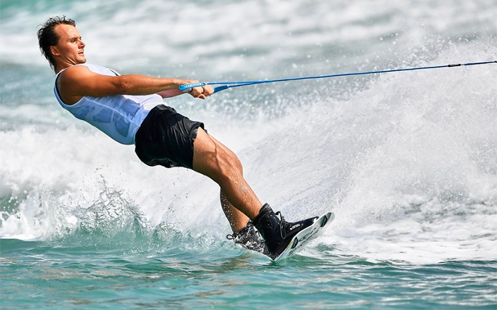 A person in a white tank top and black shorts is wakeboarding in Qatar, leaning back and gripping a tow rope as waves crash and splashes surround them on the water.