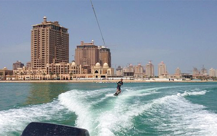 A person is wakeboarding in Qatar on turquoise water near a city shoreline with tall buildings and domed structures under a clear blue sky.