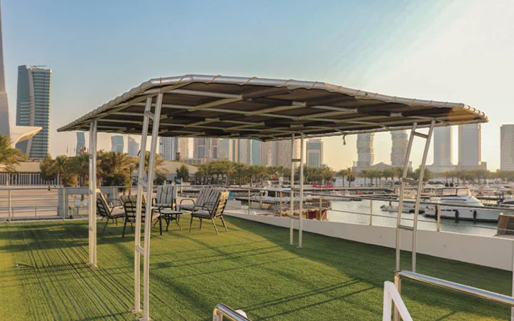 A rooftop terrace with artificial grass, outdoor chairs, and tables under a metal canopy, overlooking a marina with boats, high-rise buildings, and the Wave Private Houseboat in the background.