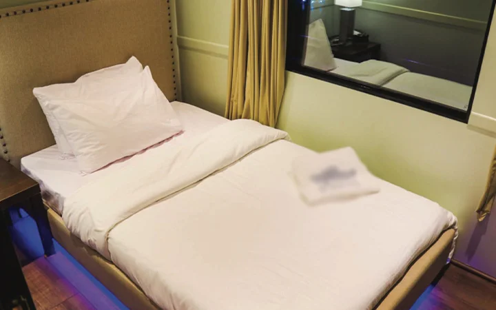 A neatly made single bed with white sheets and a pillow sits beside a window with closed curtains inside the Wave Private Houseboat. A bedside table with a lamp and a folded towel on the bed create a cozy, inviting space.