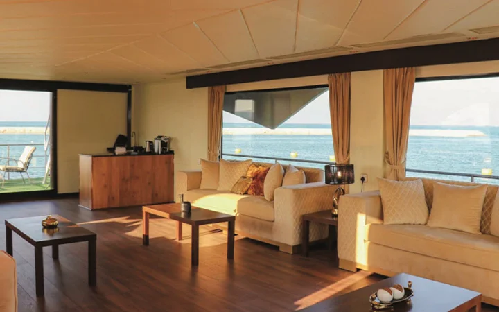 A spacious, modern living room aboard the Wave Private Houseboat with cream sofas, wooden tables, and large windows offering ocean views. Light pours in, highlighting the cozy, elegant decor and wooden floors.