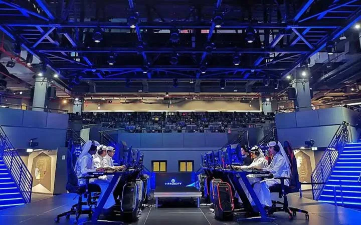 Two teams of players in traditional Middle Eastern attire compete in an esports tournament on a stage with blue lighting and elevated seating for an audience in the background.