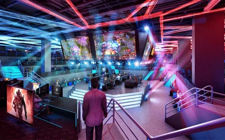 A vibrant esports arena with colorful lighting, large screens displaying a video game, teams competing on stage, and an audience watching from tiered seating. A person stands at the forefront, overlooking the scene.