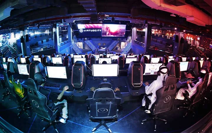 A large gaming arena with rows of people at computers, wearing headsets, facing a lit stage with big screens displaying “Virtuocity.” The space is modern, colorful, and filled with gaming chairs.