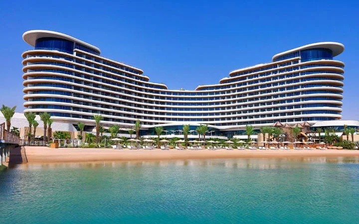 A modern, curved luxury hotel—Waldorf Astoria Beach—boasts multiple floors and balconies overlooking a sandy beach and clear blue water, with palm trees and lounge chairs lining the shore under a bright blue sky.
