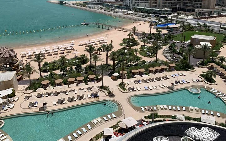 Aerial view of Waldorf Astoria Beach resort with multiple pools, palm trees, sun loungers, and umbrellas, overlooking a sandy beach and turquoise sea. People are relaxing by the pools and on the beach.