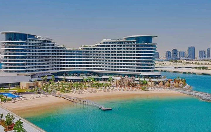 A large modern Waldorf Astoria Beach hotel with curved architecture, multiple balconies, a sandy beach with sun loungers, and turquoise water in the foreground. City skyscrapers are visible in the distant background under a clear sky.