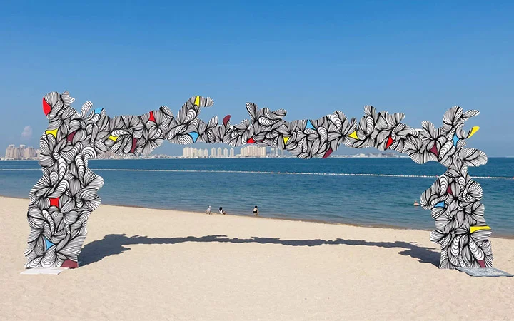 A large, abstract arch covered in black-and-white floral patterns with touches of yellow and red stands on the sandy shore of West Bay Beach, with a calm blue sea and distant city buildings forming the backdrop.