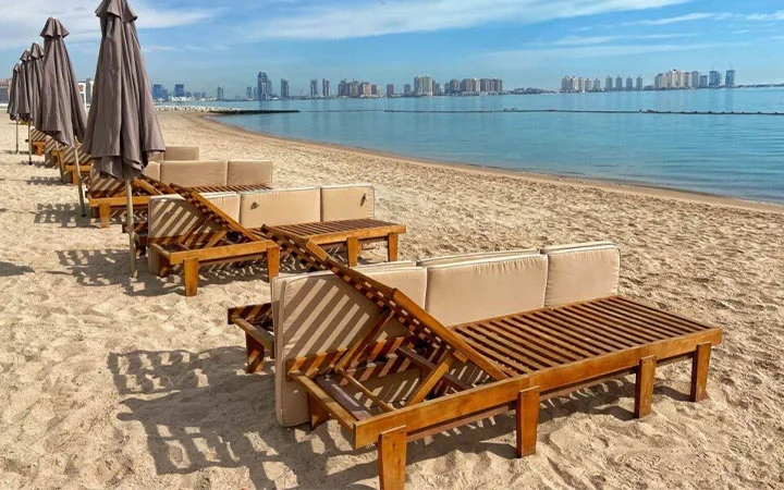 Wooden lounge chairs with beige cushions and closed umbrellas line the sandy shores of West Bay Beach, facing calm water. A distant city skyline is visible across the water under a blue sky with light clouds.