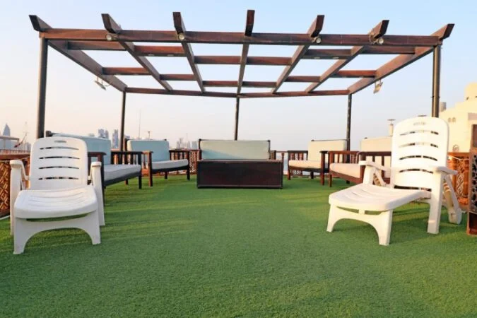Outdoor rooftop seating area with artificial grass flooring, white plastic lounge chairs, wooden benches with cushions, and a wooden pergola overhead—perfect for a クルー限定 日本航空 サンセットクルーズ under the clear sky.