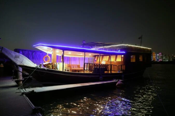 A wooden boat with glowing blue and white lights, part of a special サンセットクルーズ パッケージ, is docked at night. Its warmly lit interior reflects on the water, with a distant city skyline shining in the background.