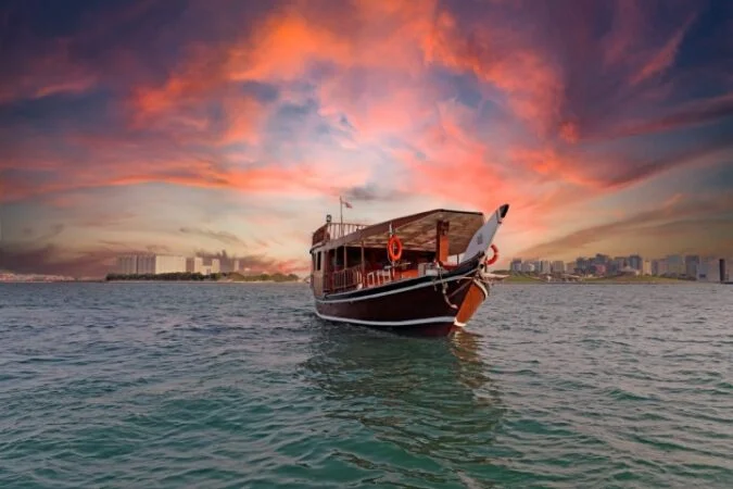 A traditional wooden boat floats on calm water at sunset, with a dramatic orange and purple sky and a distant city skyline—perfect for a serene サンセットクルーズ experience.