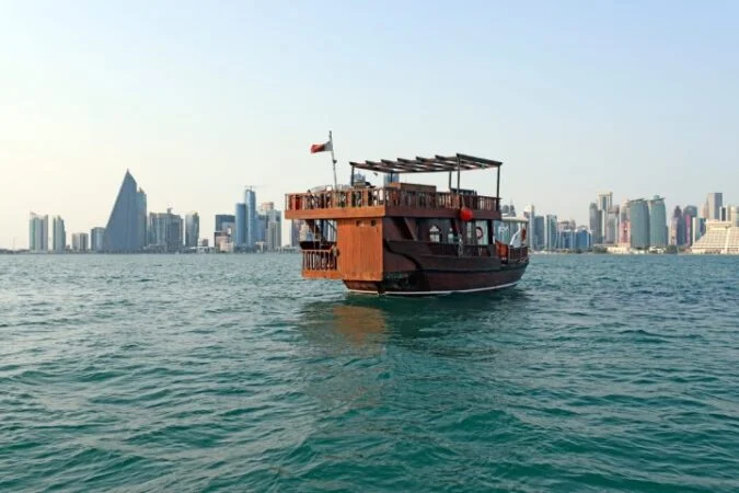 A traditional wooden dhow boat floats on blue water as the modern skyline rises behind, perfect for a サンセットクルーズ under a clear sky.